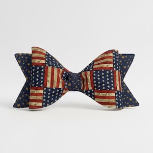 May include: A patriotic bow tie featuring a patchwork design of the American flag. The bow tie has a navy blue background with gold stars on the ends. The center of the bow tie displays the red and white stripes and blue star field of the American flag.