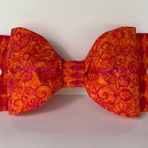 May include: A large, orange and pink bow made of fabric with a floral pattern. The bow has a row of sequins across the center.