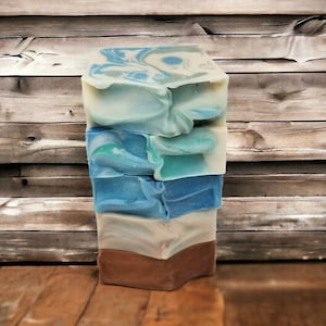 May include: A stack of four handmade soap bars with a variety of colors and patterns. The soap bars are layered on top of each other, with the top bar being a light blue and white swirl, the second bar being a light blue and green swirl, the third bar being a dark blue swirl, and the bottom bar being a brown and white swirl.