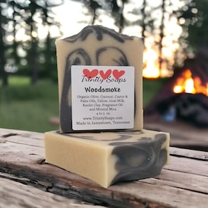 May include: A bar of handmade soap with a black and white swirl design. The soap is labeled "Trinity Soaps Woodsmoke" and lists the ingredients: Organic Olive, Coconut, Castor & Palm Oils, Tallow, Goat Milk, Kaolin Clay, Fragrance Oil and Mineral Mica. The soap is 4 to 5 ounces and made in Jamestown, Tennessee.