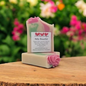 May include: Two bars of handmade soap with a floral design. The soap is white with pink and green swirls and a pink rose design. The label reads "Trinity Soaps Hello Beautiful" and lists ingredients.