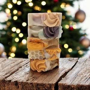 May include: A stack of handmade soaps with decorative frosting-like designs. The soaps are various colors, including beige, yellow, and purple. The soaps are stacked on a rustic wooden surface, with a blurred Christmas tree in the background.