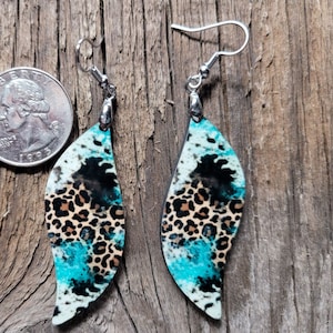 May include: A pair of dangle earrings with a teal, black, and brown leopard print design. The earrings are made of metal and have a silver hook.