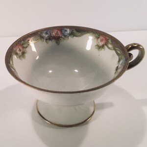 May include: A white teacup with a gold rim and floral design. The cup has a pedestal base and a handle.