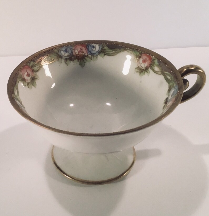 May include: A white teacup with a gold rim and floral design. The cup has a pedestal base and a handle.