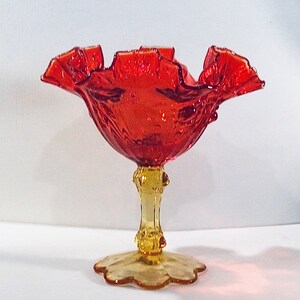 May include: A red glass candy dish with a ruffled edge and a yellow glass pedestal base.