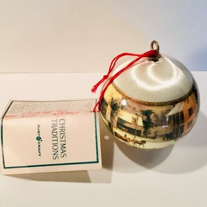 May include: A round Christmas ornament with a painted landscape scene. The ornament has a white top and a gold-colored metal loop for hanging. The tag reads "Christmas Traditions" and "Rust Craft".