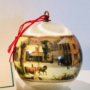 May include: A round Christmas ornament with a winter scene featuring a barn, trees, and animals. The ornament has a cream-colored base and top with a gold trim. A red string is attached for hanging.