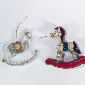 Puede incluir: Dos adornos decorativos de caballo balancín. Uno es transparente esmerilado con detalles dorados, el otro es color crema con base roja y crin marrón. Ambos tienen colgadores dorados para el árbol de Navidad.