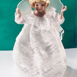 May include: A white porcelain angel figurine with blonde hair and a white lace dress. The angel has wings and is holding its hands up in a prayer position.