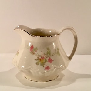 May include: A white ceramic creamer with a gold rim and a floral design. The creamer has a scalloped edge and a handle.