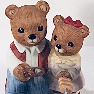 May include: A pair of brown ceramic teddy bears, one wearing a red and white vest and blue jeans, the other wearing a white dress with a yellow bow and red shoes.