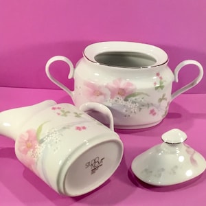 Peut inclure: Un ensemble de crémier et de sucrier en porcelaine blanche avec un motif floral. Le crémier a une poignée et un bec verseur. Le sucrier a un couvercle et deux poignées. L'ensemble est marqué "Silko Nova" au fond.