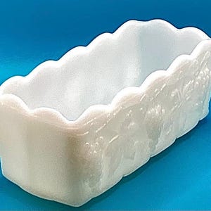 Puede incluir: Un plato rectangular de vidrio opalino blanco con un borde festoneado. El lado del plato presenta un diseño floral en relieve. El plato es probablemente para servir o exhibir.