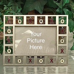 Puede incluir: Un marco de fotos plateado con un paspartú blanco y un inserto de vidrio transparente. El marco está decorado con pequeños amuletos que deletrean "XOXO". El marco tiene un lugar para una foto con el texto "Your Picture Here" impreso debajo del vidrio.