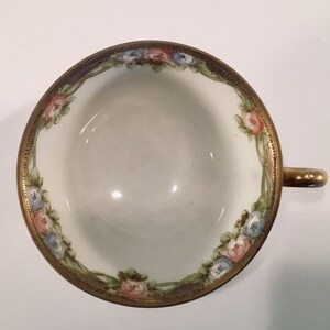 May include: A white porcelain teacup with a gold rim and floral design. The cup features pink, blue, and green flowers with gold accents.