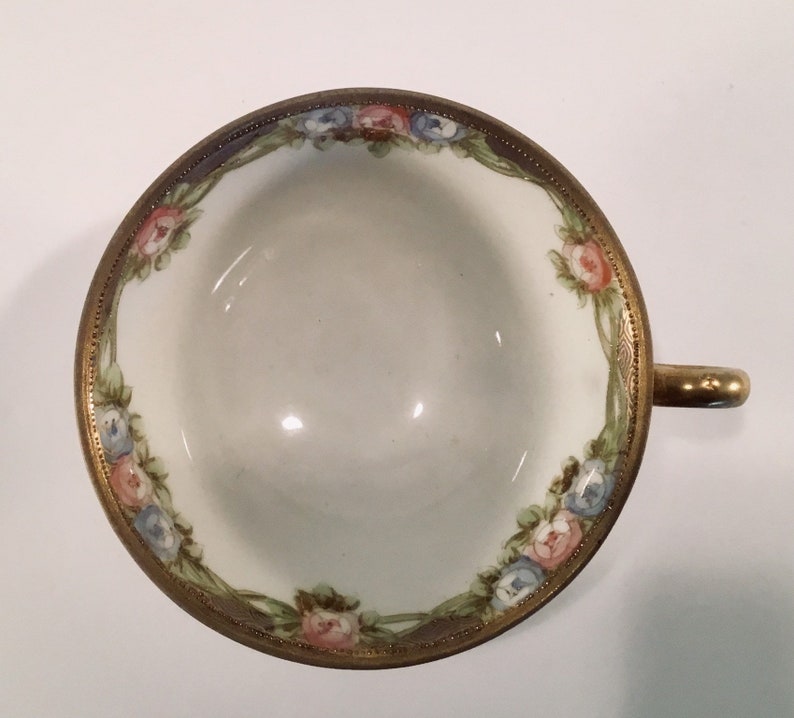 May include: A white porcelain teacup with a gold rim and floral design. The cup features pink, blue, and green flowers with gold accents.