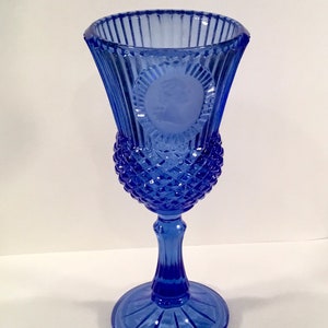 May include: A cobalt blue glass goblet with a detailed design. The goblet features a textured, diamond-patterned lower bowl, a fluted stem, and a wide, ribbed rim. A circular cameo is featured on the goblet. The goblet is a decorative piece.