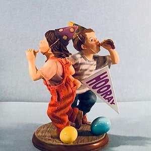 Puede incluir: Figura de dos niños celebrando. Uno lleva un mono naranja, el otro una camiseta a rayas. Ambos llevan gorros de fiesta y soplan matasuegras. Una pancarta dice "HOORAY". Un huevo amarillo y otro azul están en la base.