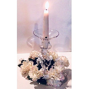 Puede incluir: Un candelero de vidrio transparente con una vela blanca encendida en su interior. El candelero está rodeado por un anillo de claveles blancos.