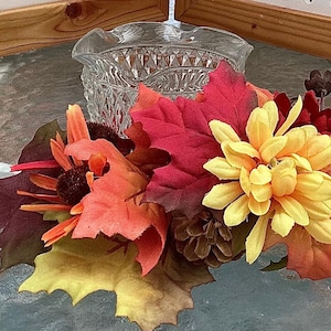 May include: A clear, cut-glass bowl with a ruffled edge sits atop a colorful arrangement of fall foliage. The arrangement includes red, orange, and yellow leaves, along with yellow flowers and a pine cone.