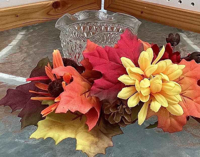 May include: A clear, cut-glass bowl with a ruffled edge sits atop a colorful arrangement of fall foliage. The arrangement includes red, orange, and yellow leaves, along with yellow flowers and a pine cone.