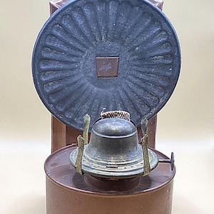 May include: Antique oil lamp with a copper-colored base and a dark gray, circular shade. The lamp features a brass burner and a decorative, rectangular top. The design is reminiscent of vintage lighting.