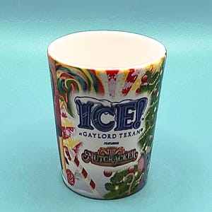 May include: A white ceramic shot glass with a colourful design. The design includes the word "ICE!" in blue, the text "GAYLORD TEXAN", and "The Nutcracker" with candy canes and Christmas trees.