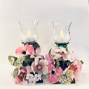 Può includere: Due portacandele in vetro con candele tealight circondate da fiori artificiali rosa e bianchi.
