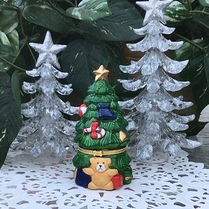 Könnte beinhalten: Eine Keramik-Weihnachtsbaum-Spieluhr mit einem Teddybären auf der Vorderseite. Der Baum ist mit Ornamenten, Zuckerstangen und einem Strumpf verziert. Die Spieluhr ist von zwei weißen Glitzer-Weihnachtsbäumen umgeben.