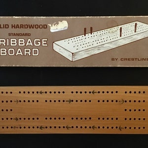 Op de afbeelding: Een houten cribbage-bord in de originele doos. De doos is bruin met de woorden "SOLID HARDWOOD STANDARD CRIBBAGE BOARD BY CRESTLINE" erop gedrukt. Het bord is gemaakt van massief hardhout en heeft een natuurlijke houtkleur.