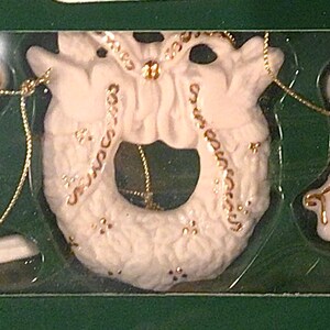 Könnte beinhalten: Set aus drei weißen Porzellan-Weihnachtsanhängern. Die Anhänger sind ein Schlitten, ein Kranz und ein Engel. Jeder Anhänger hat goldene Akzente.
