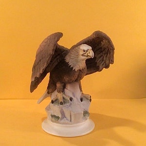 Könnte beinhalten: Eine Porzellanfigur eines Weißkopfseeadlers mit braunen und weißen Federn, der auf einem Felsen sitzt. Der Adler hat seine Flügel ausgebreitet und den Kopf zur Seite gedreht. Die Figur steht auf einem weißen Sockel.