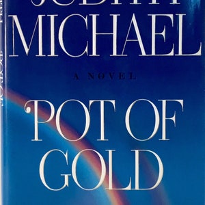 Puede incluir: Una cubierta de libro azul con el título "Pot of Gold" en texto blanco. El nombre del autor, Judith Michael, está escrito en texto blanco sobre el título. La cubierta del libro también presenta un arcoíris y un barco en la distancia.