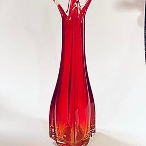 Può includere: Un vaso alto e sottile in vetro rosso, con la parte superiore svasata e la base a punta. Il vaso ha un bordo in vetro trasparente, quasi bianco, e una leggera sfumatura ambrata sul fondo.