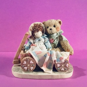 May include: A figurine of a teddy bear and a doll sitting in a wagon. The wagon is decorated with hearts and the doll is wearing a blue dress. The teddy bear is wearing a brown and white striped shirt and a red bow tie.