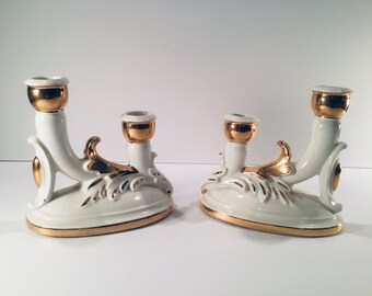 May include: A pair of white ceramic candle holders with gold accents. The holders have a unique design with a curved base and a single candle holder on each side.