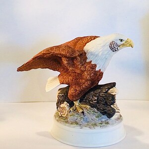 Könnte beinhalten: Eine Porzellanfigur eines Weißkopfseeadlers mit braunen und weißen Federn, der auf einem Baumstumpf sitzt. Der Adler hat einen weißen Kopf und einen gelben Schnabel. Der Sockel der Figur ist weiß und hat eine braune und grüne bemalte Szene.