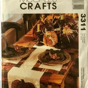 May include: A McCall's Crafts sewing pattern for a table runner with a fall theme. The pattern includes instructions for making a table runner with a patchwork design featuring plaid fabric and a pumpkin applique. The pattern number is 3311.