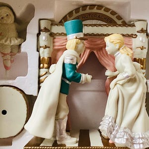 May include: A decorative figurine set depicting characters from "The Nutcracker" ballet. The set includes a male figure in a teal jacket and white cape, and a female figure in a white dress. A small ballerina and a drum are also visible.