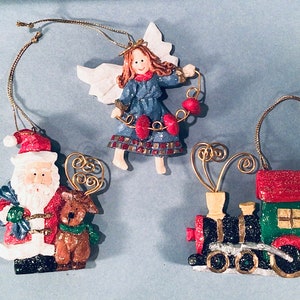 May include: Three Christmas tree decorations: a Santa Claus figure with a reindeer, an angel, and a train. The Santa ornament has a red hat and coat, the angel wears a blue dress, and the train is black and green. All have gold hanging strings.