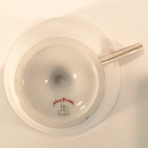 May include: The bottom of a white teacup with a silver handle. The cup has a red and gold hand-painted design that says "Hand Painted" and "R & C Nippon".