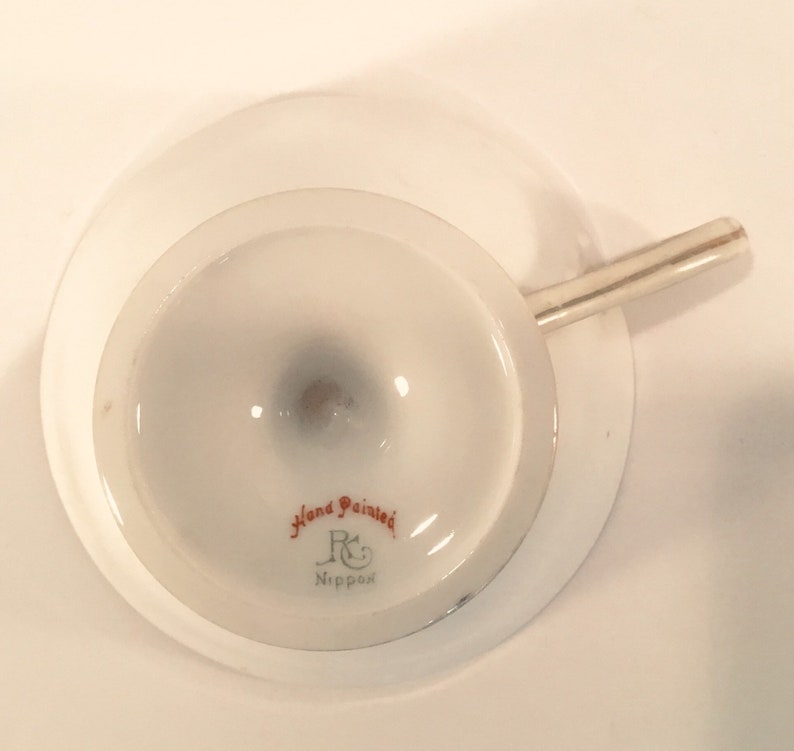 May include: The bottom of a white teacup with a silver handle. The cup has a red and gold hand-painted design that says "Hand Painted" and "R & C Nippon".