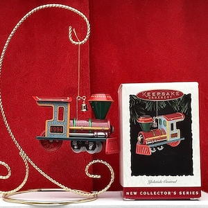 May include: A red, green and blue toy train ornament with a gold bell hanging from the top. The ornament is hanging from a gold stand. The box for the ornament says "Keepsake Ornament" and "Yuletide Central" with a red and green Christmas tree design. The box also says "New Collector's Series".