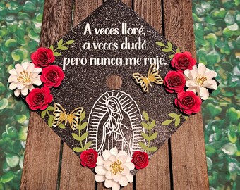 May include: A black graduation cap decorated with red and white paper flowers, green leaves, and gold butterflies. The cap has a white silhouette of the Virgin Mary and the text "A veces lloré, a veces dudé pero nunca me rajé."