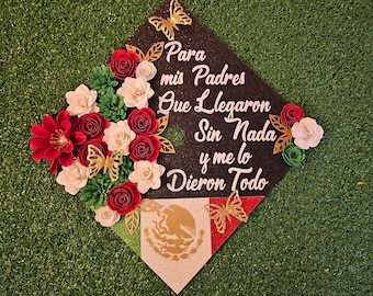 Puede incluir: Un gorro de graduación negro decorado con flores de papel rojo y blanco, mariposas doradas y una bandera mexicana verde, blanca y roja. El gorro tiene el texto "Para mis Padres Que Llegaron Sin Nada y me lo Dieron Todo" escrito en él.