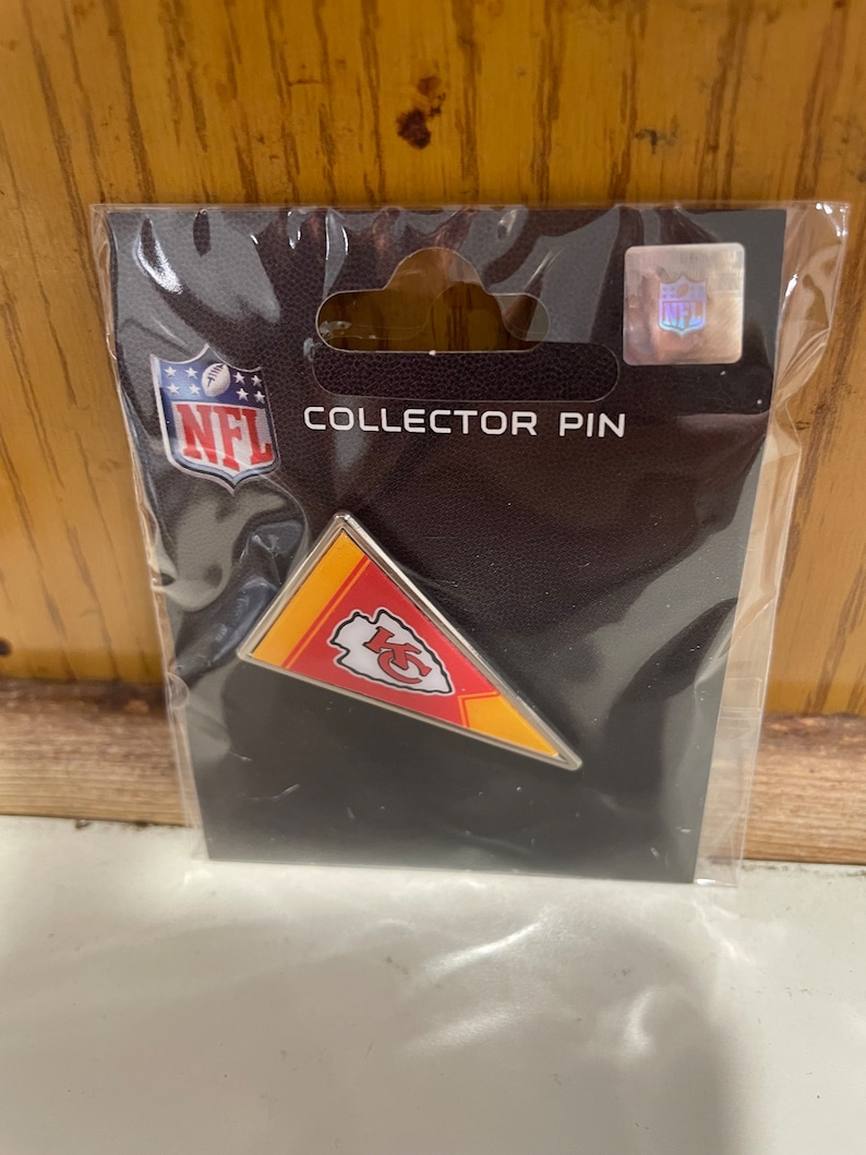 Może przedstawiać: Przypinka kolekcjonerska z logo Kansas City Chiefs, zapakowana w przezroczysty plastikowy rękaw. Przypinka ma kształt proporczyka w kolorach czerwonym, ż&oacute;łtym i białym. Opakowanie zawiera logo NFL i napis "COLLECTOR PIN".