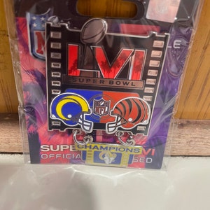 May include: A Super Bowl LVI commemorative pin set featuring the Los Angeles Rams and Cincinnati Bengals helmets. The pins are silver with blue, red, and yellow accents. The set includes a film strip with the text "Super Bowl LVI" and the NFL logo.