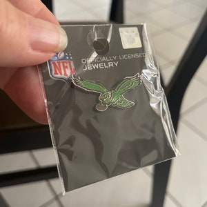 May include: Officially licensed NFL jewelry featuring a green Philadelphia Eagles pin on a black card with the NFL logo.