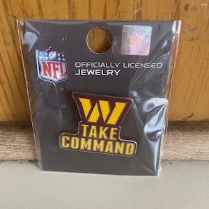 May include: A black and gold NFL pin with the Washington Commanders logo and the text "TAKE COMMAND". The pin is officially licensed NFL jewelry.
